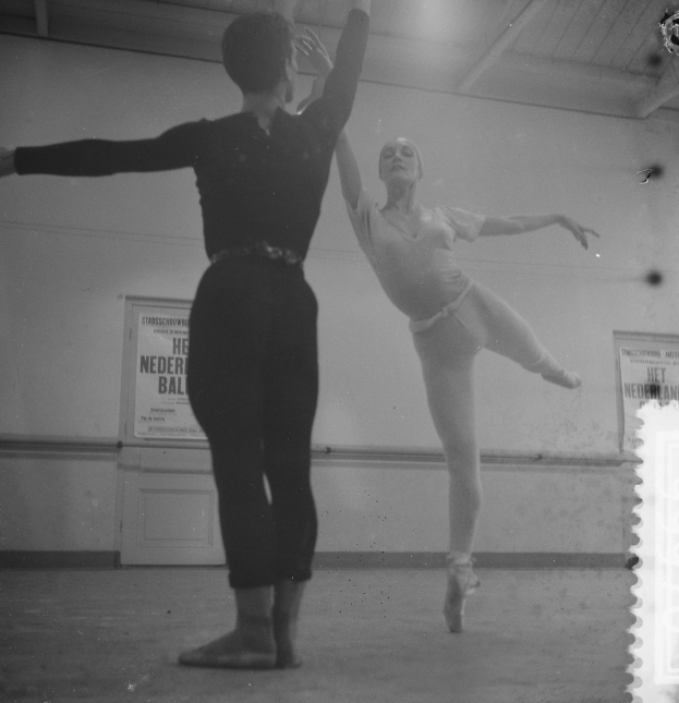 Ein Schwarz-Weiß-Foto von einem Mann und einer Frau, die in einem Studio tanzen, wobei die Frau in der Luft ist und eine mit Postern bedeckte Wand im Hintergrund zu sehen ist, die von oben beleuchtet wird.