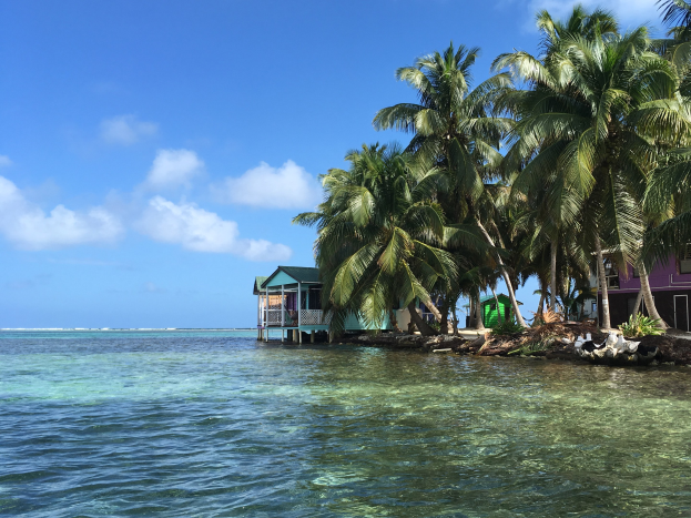 Kleiner Insel mit Palmen, klarem blauem Wasser, weißen Wolken am Himmel und Häusern auf der rechten Seite.