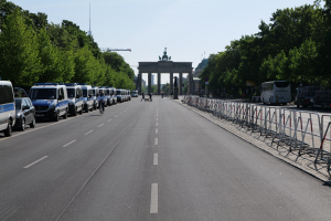Lange Reihe von Polizeiwagen auf der Seite einer Straße vor dem Brandenburger Tor in Berlin, Deutschland, mit Menschen auf Fahrrädern und in der Nähe stehenden, Barrieren, Bäumen, einem Bogen mit Statuen im Hintergrund und sichtbarem Himmel.