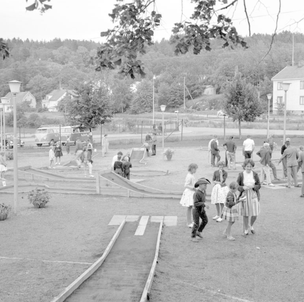 Schwarz-weißes Bild von Menschen, die auf einem Parkplatz mit Grünflächen, Laternen, geparkten Fahrzeugen und Gebäuden im Hintergrund Minigolf spielen.