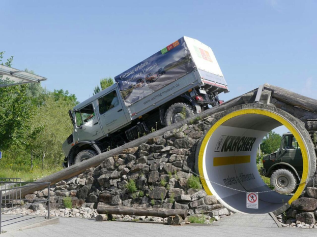 Ein Lastwagen fährt durch einen Stein- und Holztunnel in einem Park, mit einem Schild, Geländern und einem Schuppen links und Bäumen und einem blauen Himmel im Hintergrund.
