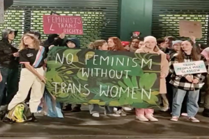Gruppe von Menschen mit einem Banner 'Kein Feminismus ohne Trans-Frauen' und Schildern in öffentlichem Raum.