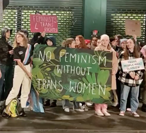 Gruppe von Menschen mit einem Banner 'Kein Feminismus ohne Trans-Frauen' und Schildern in öffentlichem Raum.