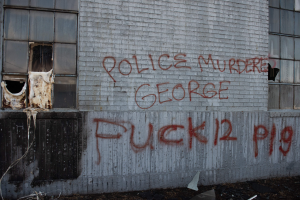 Schmiererei an einer Hauswand mit der Aufschrift "Polizeimord George Fuck 12 P19" mit Fenstern und verstreuten Gegenständen davor.