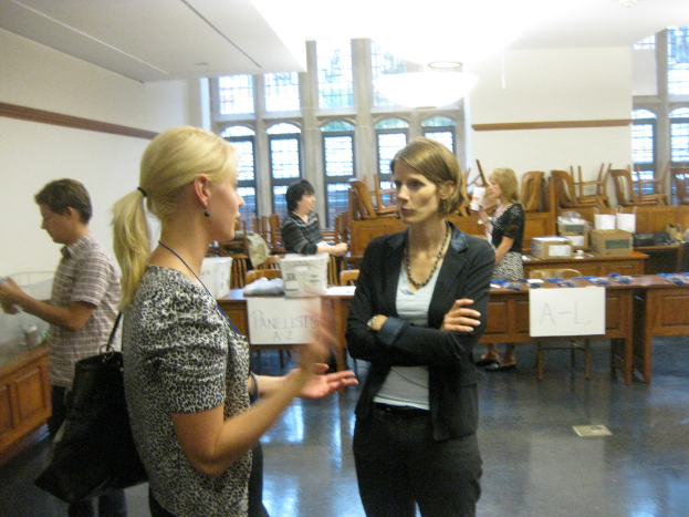 Zwei Frauen stehen und sprechen miteinander, wobei die Frau links eine Handtasche trägt. Im Hintergrund hält ein Mann ein Objekt, und zwei weitere Frauen stehen hinter einer Bank mit beschrifteten Gegenständen.