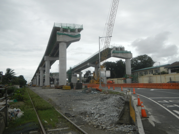 Baustelle mit Brücke im Hintergrund, Straße mit Absperrbaken, Bahnschiene, Steine, Gras, Bäume, Gebäude und bewölkter Himmel.