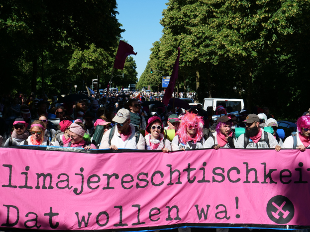 Eine Gruppe von Menschen marschiert stra√enabwärts und hölt eine rosafarbene "Women's March in Berlin"-Fahne, einige tragen Mützen und Schutzbrillen und andere halten Fahnen, vor dem Hintergrund von Bäumen, Fahrzeugen und Himmel.