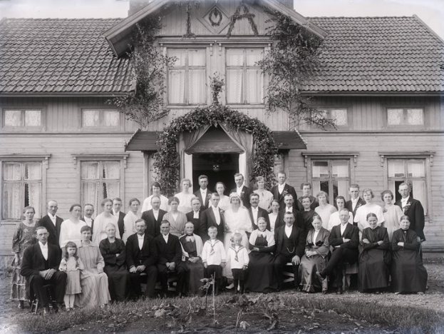 Schwarze-weisse-Foto-einer-großen-Gruppe-Menschen-vor-einem-Haus-mit-grüner-Umgebung-und-sichtbarem-Himmel.