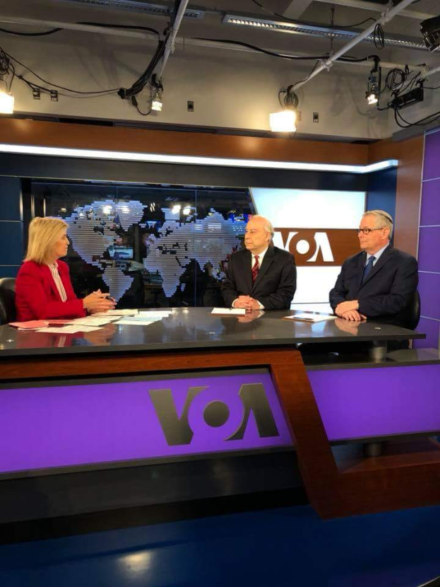 Drei Personen, zwei Männer in Blazern und Krawatten und eine Frau in einem roten Kleid sitzen an einem Schreibtisch mit Papieren und Gegenständen, diskutieren vor einem Fernseher mit einem Bildschirm und Lampen im Hintergrund.