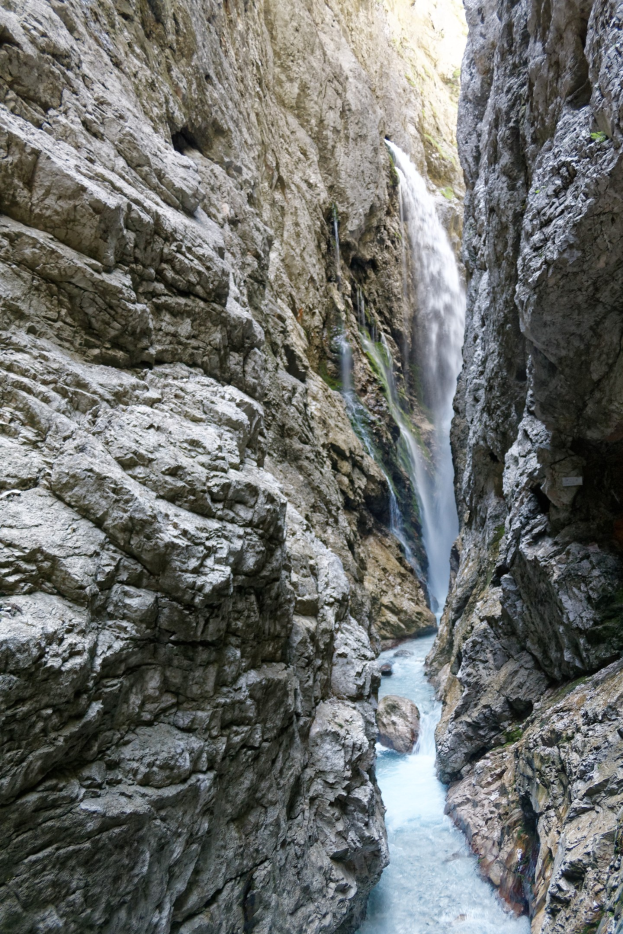 Ein kleiner Wasserfall stürzt sich über zerklüftete Felsen in einem steinigen Tal, umgeben von saftig grünen Hügeln unter strahlendem Sonnenschein.