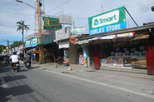 Eine belebte Straße mit Motorrädern, Autos, Gebäuden, Straßenschildern, Strommasten, Stromleitungen, Straßenlaternen, Bäumen und einem bewölkten Himmel.