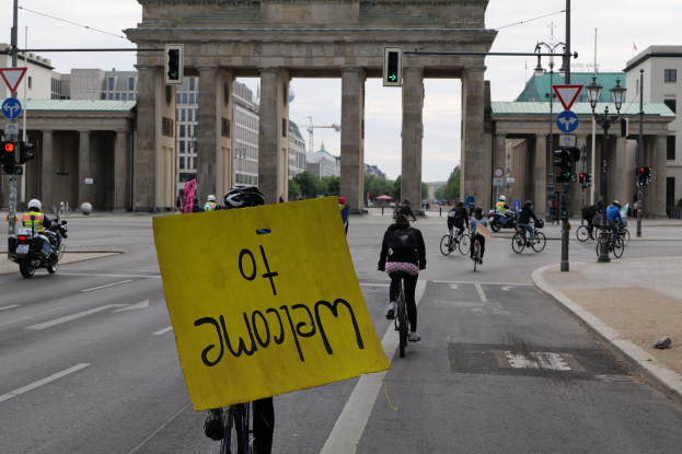 Eine Gruppe von Menschen mit Helmen fährt Fahrräder auf einer Straße vor dem Brandenburger Tor in Berlin, Deutschland, vorbei, wobei eine Person ein gelbes Schild hält, Laternenpfähle, Verkehrszeichen, Gebäude, Bäume und einen klaren blauen Himmel im Hintergrund.