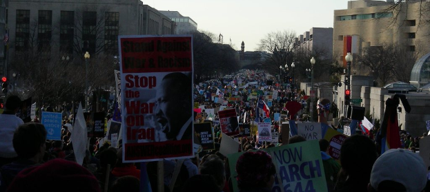 Eine Gruppe von Menschen, die auf der Straße protestieren und Plakate halten, mit Gebäuden, Bäumen und Laternenmästen im Hintergrund unter einem klaren Himmel.