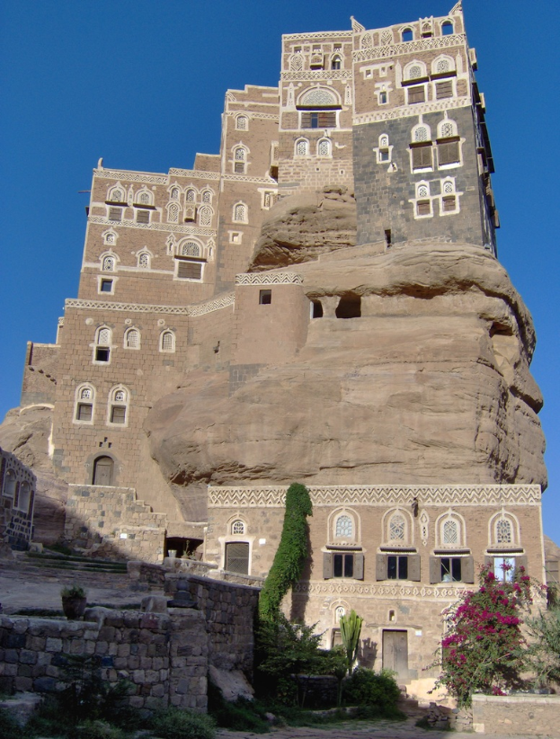 Eine Burg mit Fenstern und Türen, umgeben von Pflanzen, unter einem blauen Himmel.