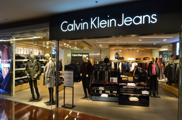 Innenansicht eines Calvin Klein Jeans-Geschäfts in einem Einkaufszentrum, das Mannequins in Kleidungsauslagen, hängende Kleidung, eine Werbetafel, einen digitalen Bildschirm und eine Deckenbeleuchtung zeigt.