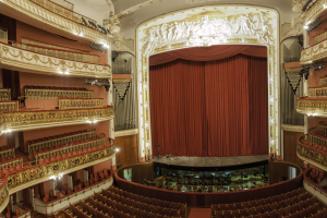 Großer Zuschauerraum mit roter Gardine, Sitzreihen, Geländern, Scheinwerfern, Wandskulpturen und minimalen Bühnenobjekten.