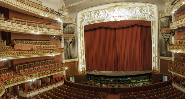 Großer Zuschauerraum mit roter Gardine, Sitzreihen, Geländern, Scheinwerfern, Wandskulpturen und minimalen Bühnenobjekten.