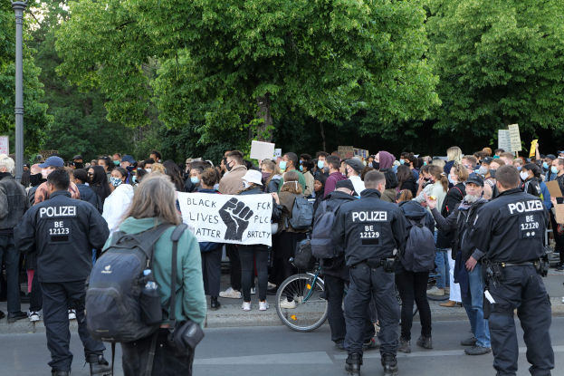 Eine große Gruppe von Menschen, die an einer Straße teilnehmen, einige halten Schilder und andere tragen Mützen und Taschen, mit einem Fahrrad im Vordergrund und Bäumen und einem Pfahl im Hintergrund.