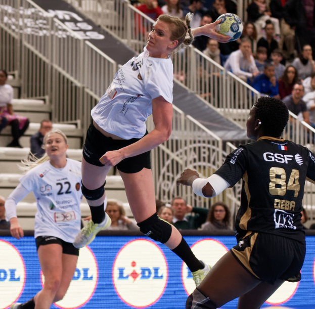 Frauen spielen Handball auf einem Feld mit einem Ball in der Luft, Zuschauern im Hintergrund und einem "Handball-Weltmeisterschaft 2019 - Tag 2"-Schild.