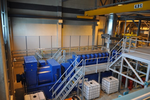 Eine Fabrikinnenszene mit Trägern, einer Treppe, Geländer, Containern, Lichtern, Röhren und Wänden.