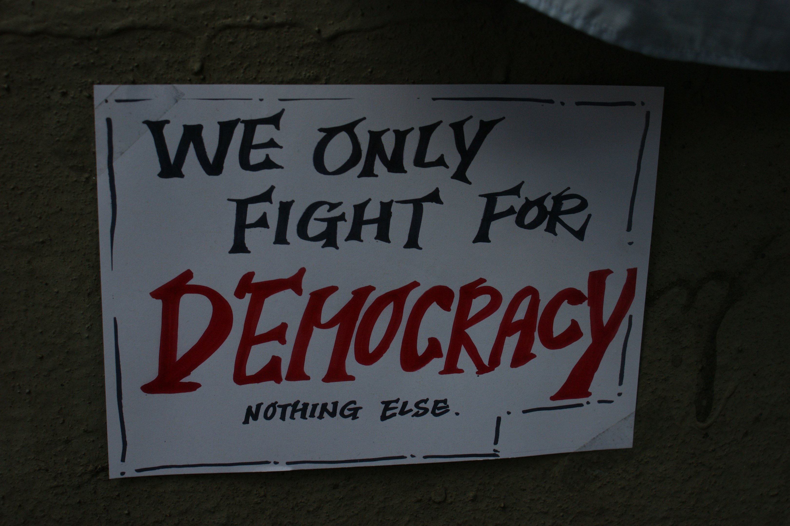 'We Only Fight for Democracy' auf weißem Blatt Papier, an einer Wand geklebt.
