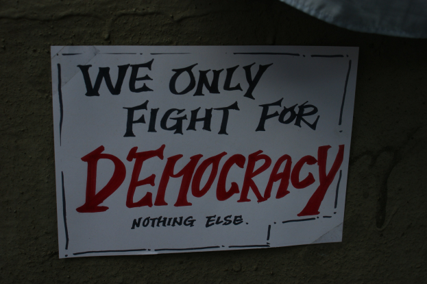 'We Only Fight for Democracy' auf weißem Blatt Papier, an einer Wand geklebt.