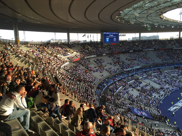 Große Menschenmenge in einem Stadion bei einem Fürballspiel mit einer Bühne, Fahnen, Masten, einem Bildschirm und dem Allianz Stadion in München, Deutschland im Hintergrund.