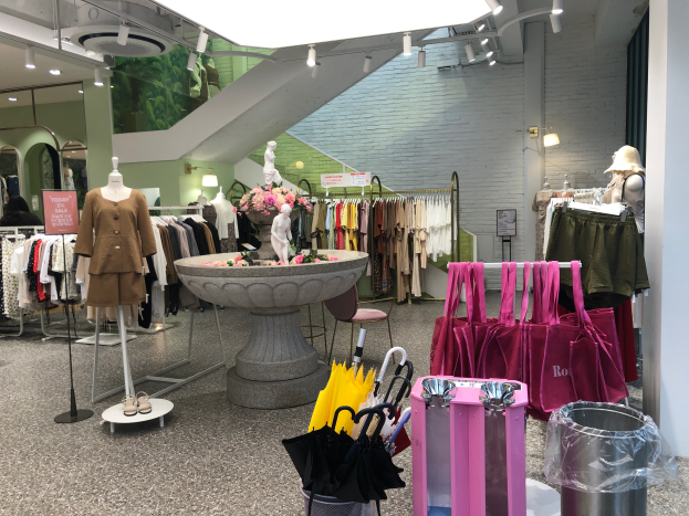 Der Innenraum eines Modegeschäfts mit Schaufensterpuppen, hängenden Kleidern, einem zentralen Brunnen, einer Blumenvase, einem Müllbehälter und Taschen.