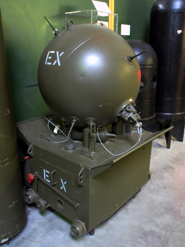 Eine große grüne Splitterbombe mit zylindrischem Metallkörper steht auf einem Tisch in der Mitte, umgeben von verschiedenen Gegenständen auf dem Boden, mit einer Wand im Hintergrund und Text am unteren Bildrand.