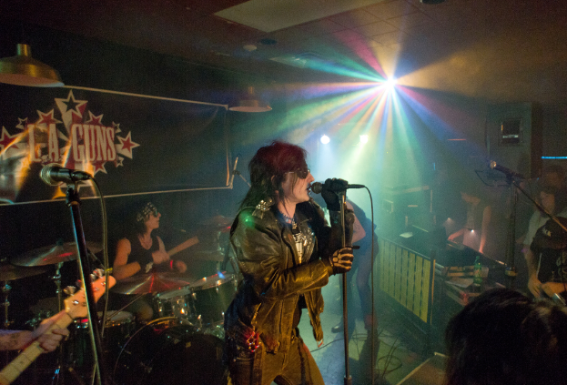 Eine Frau in einer schwarzen Jacke singt auf der Bühne in ein Mikrofon, während ein Mann mit Sonnenbrille und Stirnbinde hinter ihr ein Instrument spielt; ein 'LA Guns'-Plakat und Diskoleuchten sind im Hintergrund zu sehen.