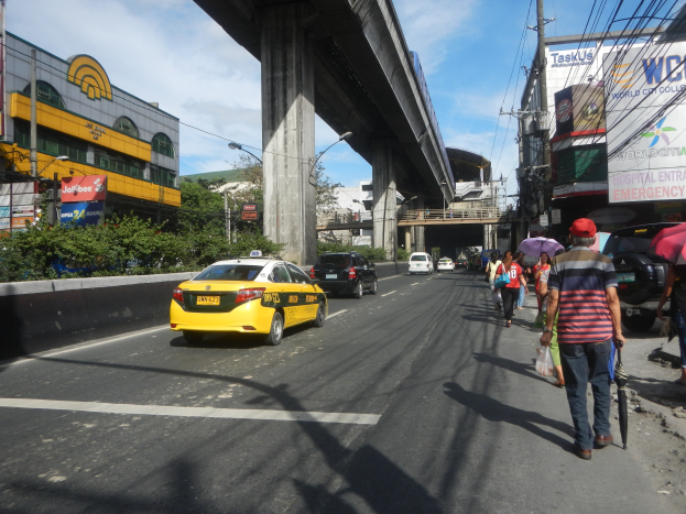 Gelbes Taxi fährt auf einer Straße mit hohen Gebäuden, Menschen gehen auf dem Bürgersteig, einige halten Regenschirme, eine Brücke im Hintergrund, bewölkter Himmel und Strommasten mit Drähten entlang der Straße.