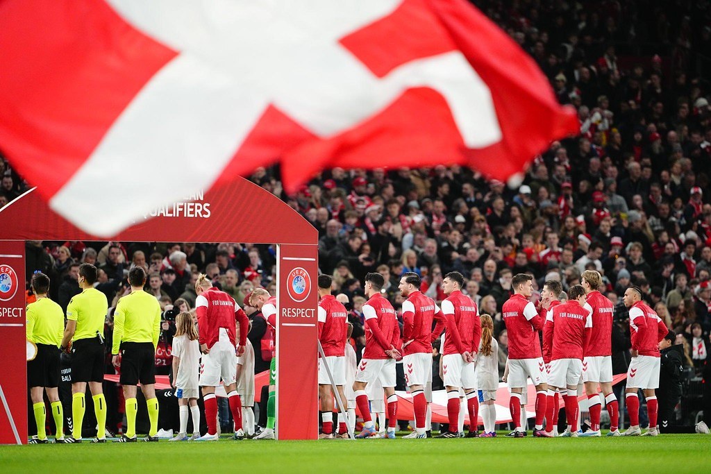 Gruppe von Menschen auf einem Fußballfeld mit einer roten und weißen Flagge im Vordergrund, einem Bogen mit der Aufschrift "Bayern München vs. Bayern München Wetten & Vorschau" im Hintergrund und einer großen Menge im Stadion.