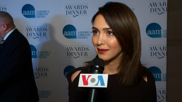 Emmy Rossum, eine Frau in einem schwarzen Kleid, hält ein Mikrofon mit einem Mann zu ihrer linken und einem Banner mit der Aufschrift "Aam Awards Dinner" im Hintergrund.