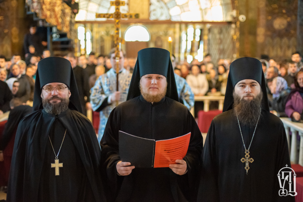 Drei Priester in schwarzen Roben und Hüten stehen vor einer Menge in einer Kirche, einer hält ein Buch, mit einem unscharfen Hintergrund und einem Wasserzeichen in der rechten unteren Ecke.