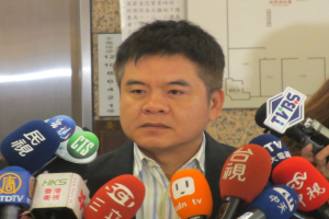 Ein Mann im Anzug und Krawatte spricht vor einer Wand mit einer Tafel zu Reportern, während eine Frau mit einer Kamera rechts steht.