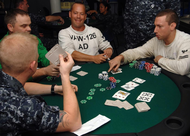 Eine Gruppe von Männern, die Poker an einem Tisch mit Münzen und Karten vor sich spielen, in einem Turnier-Setting mit einer Säule im Hintergrund.