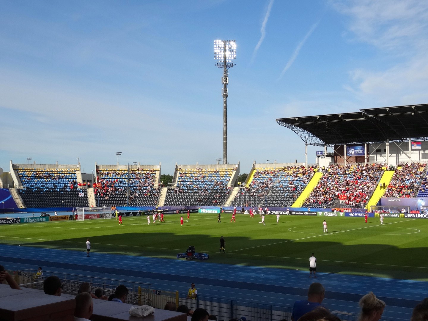 Ein Fußballspiel in einem großen, flutlichtbeleuchteten Stadion mit Zuschauern auf den Rängen und Spielern auf dem Feld unter einem sichtbaren Himmel.