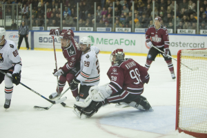 Gruppe von Menschen, die Hockey auf einem Eisstadion spielen, mit Torpfosten auf der rechten Seite, tragen Helme und halten Stöcke, Zuschauer auf den Tribünen mit Bannern im Hintergrund.