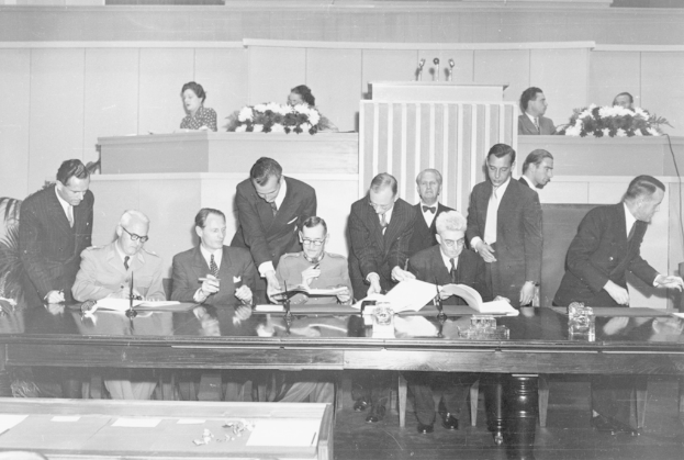 Eine Gruppe von Menschen, die auf Stühlen sitzen und andere hinter ihnen an einem Tisch mit Mikrofonen und Gläsern und einem weiteren Tisch mit Blumenbouquets und mehr Menschen stehen.