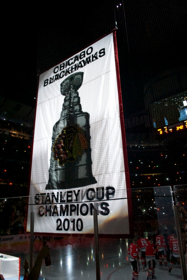 Eine nächtliche Stadionszene mit einem großen Banner in der Mitte, Zuschauern im Hintergrund, einem Glas auf dem Boden und einigen Spielern auf dem Feld, beleuchtet von Stadionlampen.