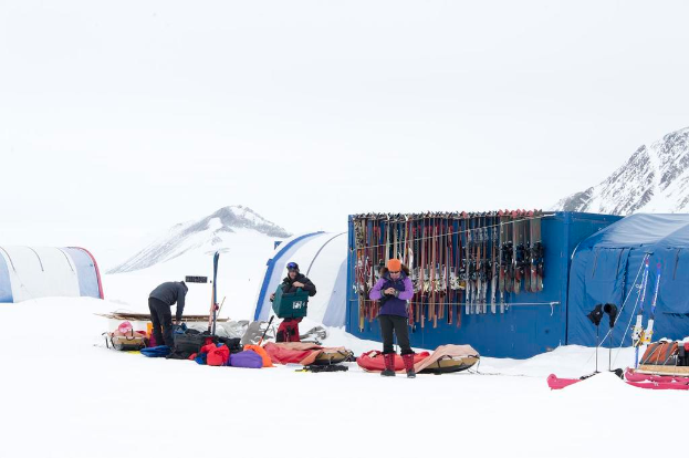 Drei Personen stehen auf einer verschneiten Landschaft mit Taschen im Hintergrund, Zelte mit Skiern darauf und schneebedeckte Hügel unter einem klaren Himmel.