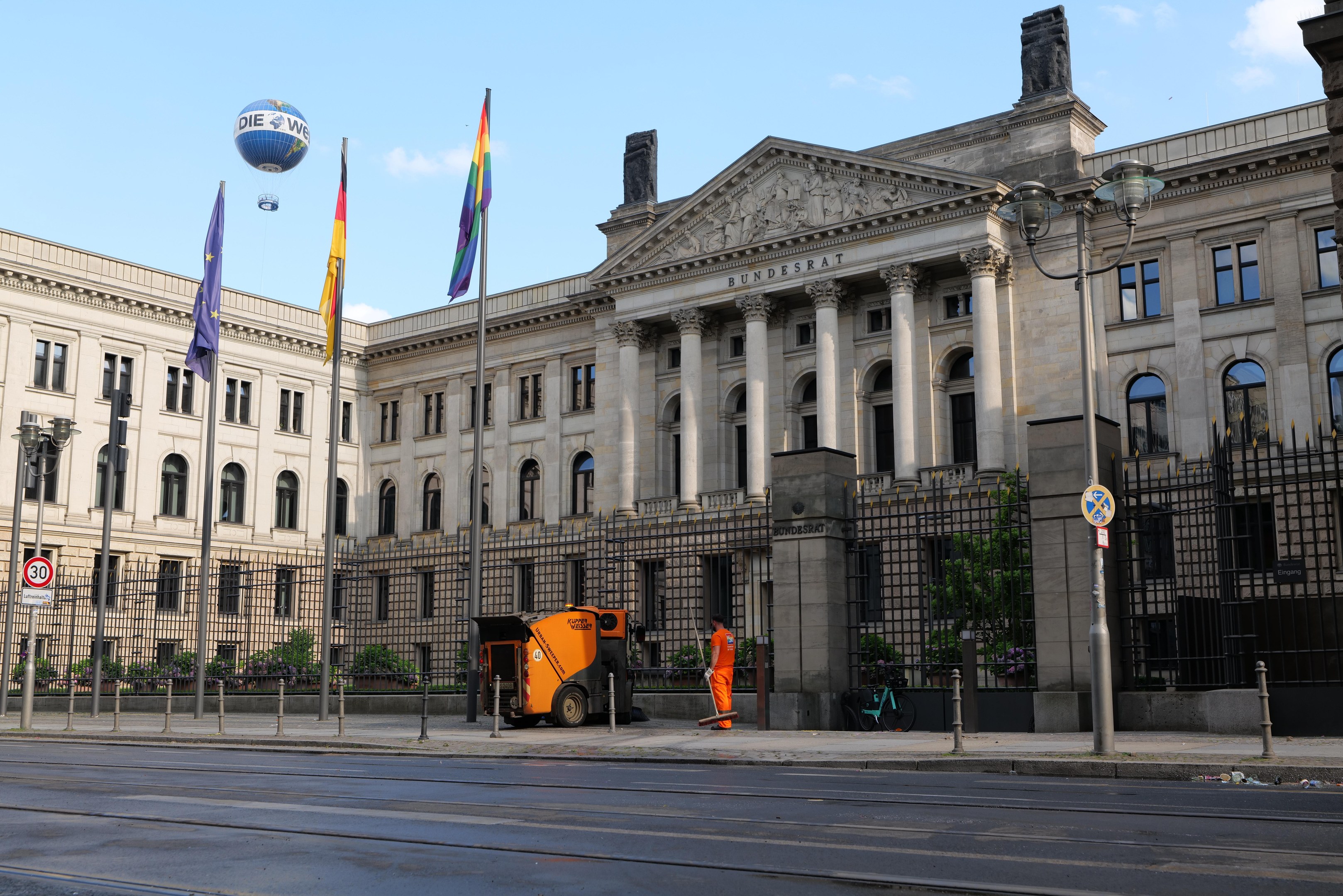 Großes beige Gebäude mit vielen Fenstern, als Bundestag in Berlin, Deutschland identifiziert, umgeben von Straßeninfrastruktur, Fußgängern, Fahrzeugen und Bäumen unter einem bewölkten Himmel.