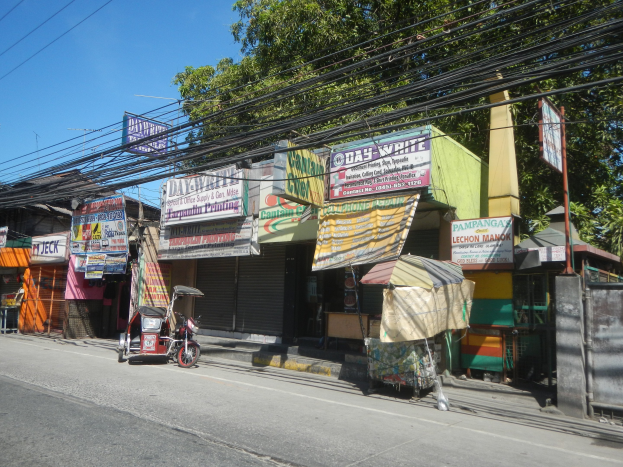 Eine Straßenszene mit einem geparkten Motorrad, Gebäuden, Geschäften, Schildern, Strommasten, Kabeln, Bäumen und einem klaren blauen Himmel.