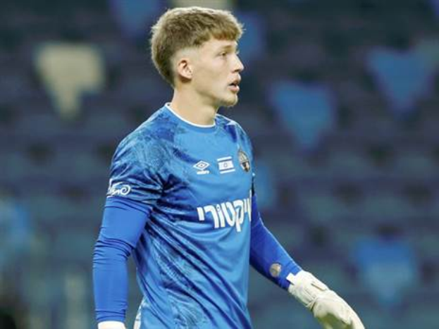 Ein junger Mann in einem blauen Torwart-Outfit und weissen Handschuhen steht auf einem Fussballfeld wahrend eines Spiels, mit einem unscharfen Hintergrund.