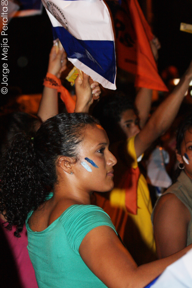 Eine Frau in einem gr√ľnen T-Shirt steht auf einer Demonstration und h√∂lt ein weißes Schild in der Hand, umgeben von vielen anderen Menschen.