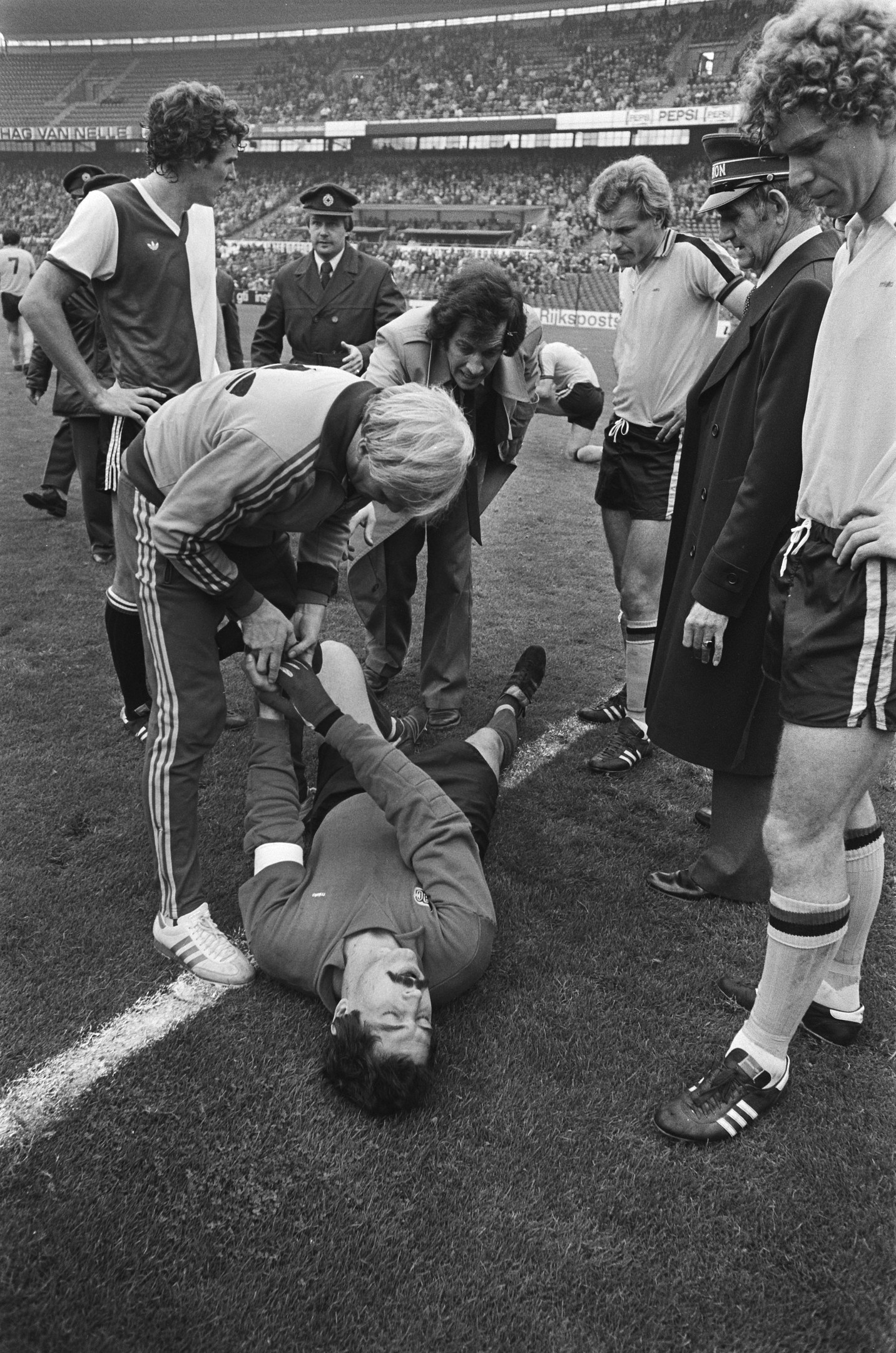 Schwarzes und weißes Bild eines Fußballspielers, der auf dem Boden liegt und von einer Gruppe Männer und einer Stadionmenge umgeben ist, wobei ein Mann zu helfen scheint.