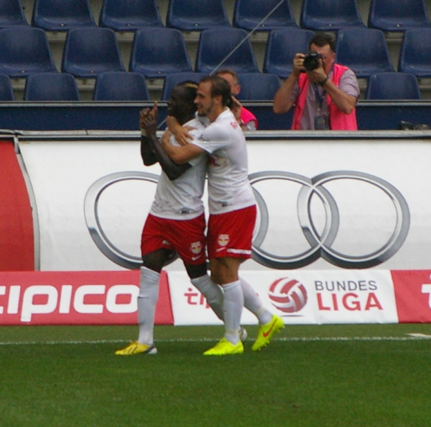 Zwei Fußballspieler feiern ein Tor auf dem Feld, umgeben von einer Gruppe von Menschen, mit einer Person, die eine Kamera hält, einem Banner mit Text, einigen Stühlen und einer Wand im Hintergrund.