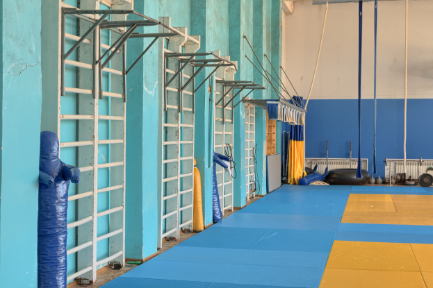 Ein Turnhalle mit blauen und gelben Matten auf dem Boden, Leitern, Stangen, Seile und andere Gegenstände, mit einer Wand im Hintergrund und einer Decke oben.
