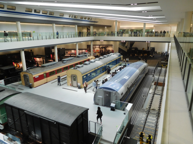 Ein großer Museumsraum mit verschiedenen Bahnen in unterschiedlichen Größen und Farben, bei dem Menschen die Ausstellungsstücke und architektonische Elemente wie Geländer, Säulen und Deckenleuchten betrachten.