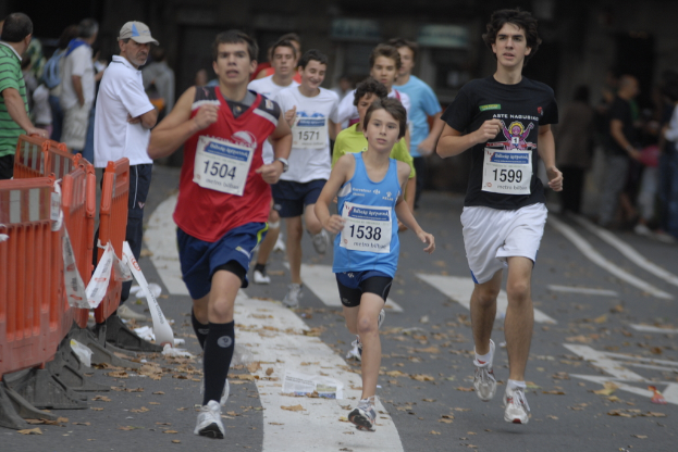 Läufer auf einer Straße mit einer orangen Absperrung auf der linken Seite, einige Zuschauer stehen und schauen zu, und nummerierte Startnummern auf den T-Shirts der Läufer.